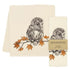 A cream-colored cotton tea towel with a detailed black-and-white owl perched on a branch with orange autumn leaves, shown alongside its paper packaging label.