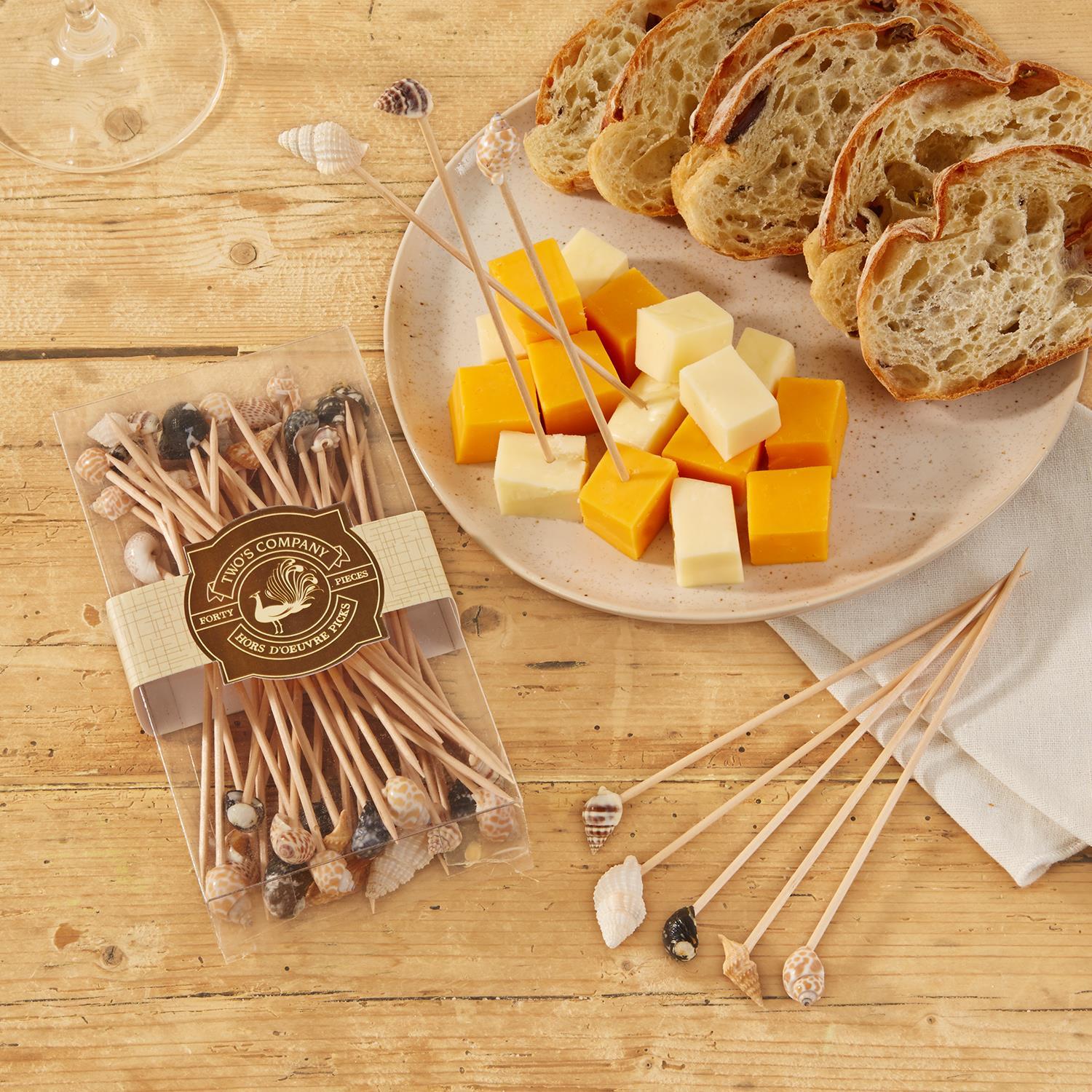 Cheese platter with seashell skewers, bread, and a box of skewers on a wood surface.