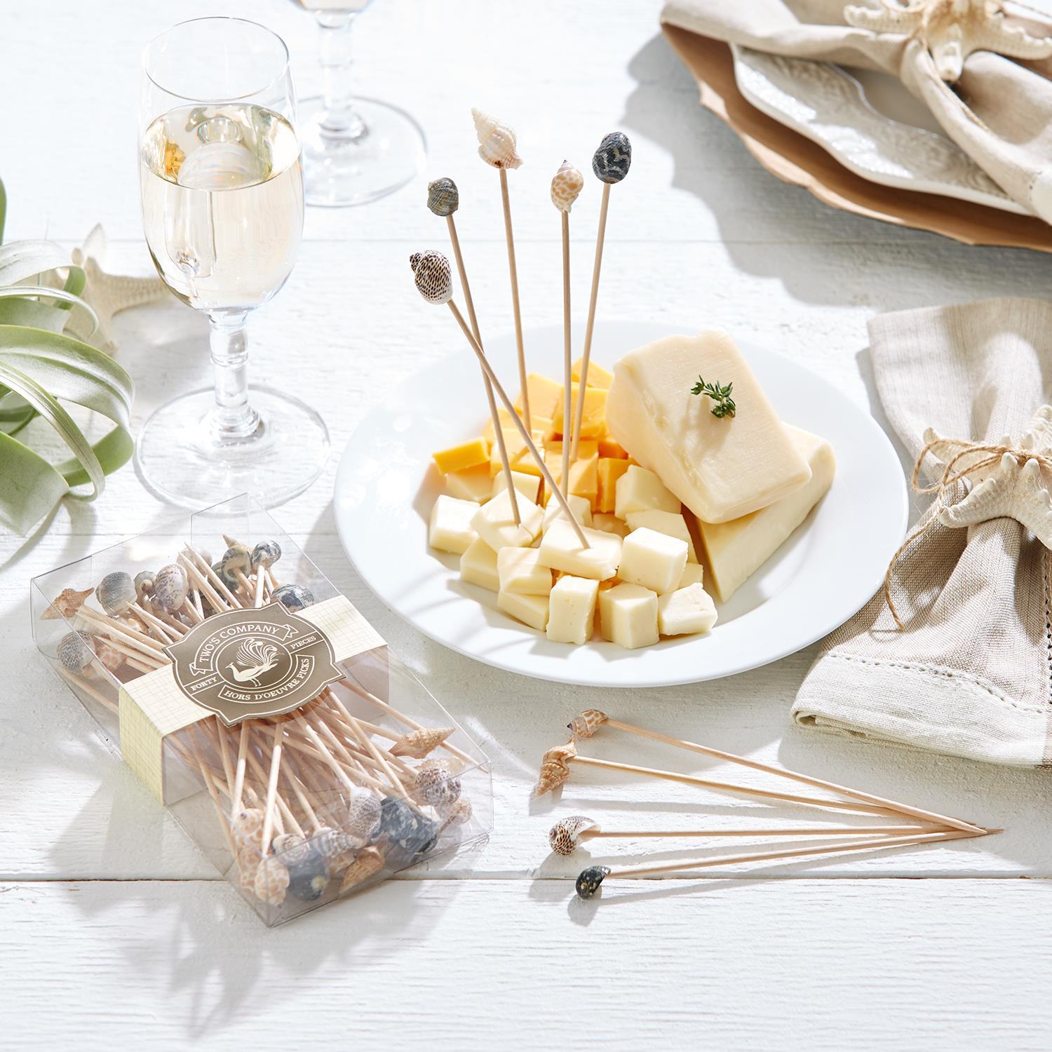 Cheese platter with seashell skewers, glasses of white wine, and a box of skewers on a white surface.