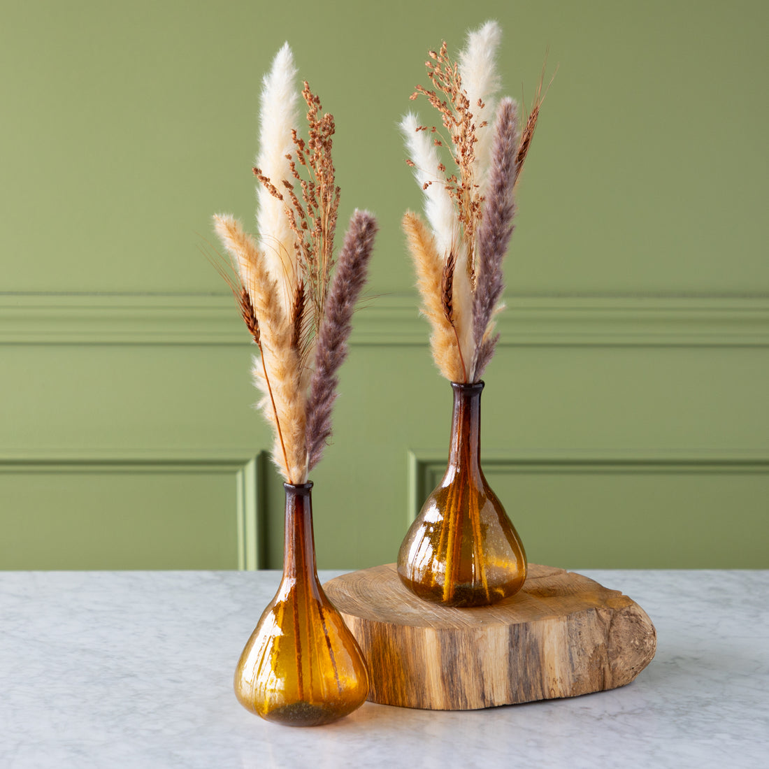 Two amber glass vases with dried pampas grass on a wooden stand against a green wall.