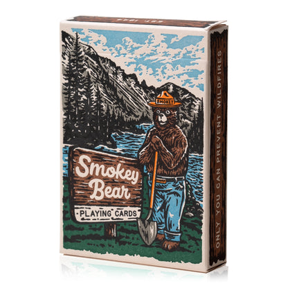 A deck of Smokey Bear Playing Cards with wildlife-themed designs, including Smokey Bear, set against a backdrop of more cards showing animal faces and fire-prevention tips. Made by Art of Play.