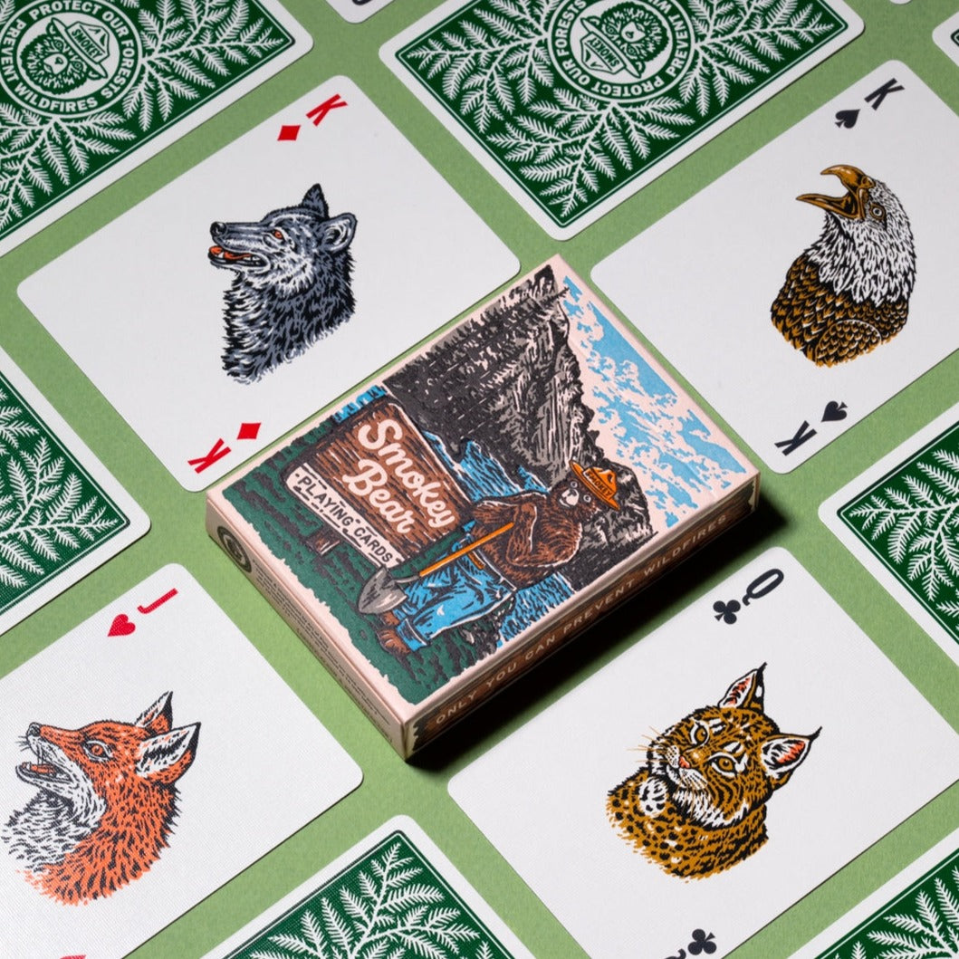 A deck of Smokey Bear Playing Cards with wildlife-themed designs, including Smokey Bear, set against a backdrop of more cards showing animal faces and fire-prevention tips. Made by Art of Play.