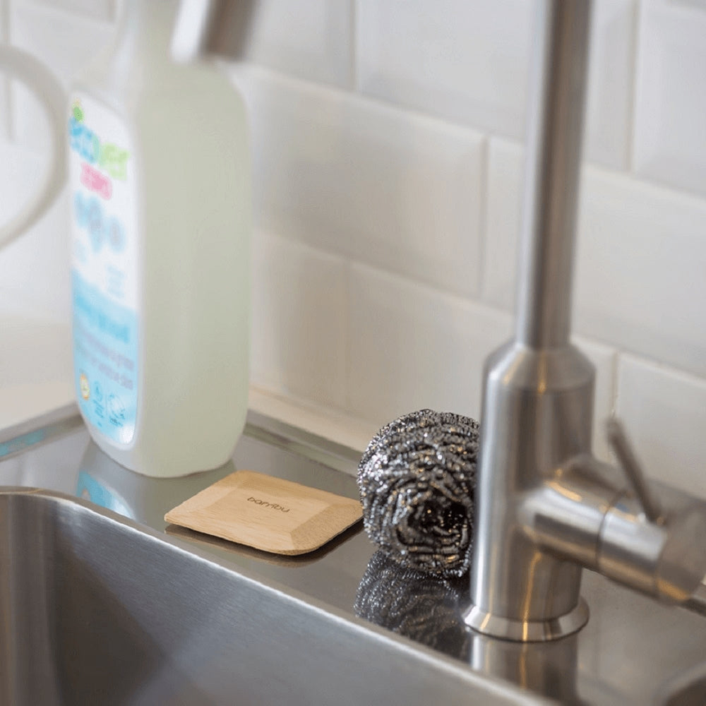 Kitchen sink with cleaning supplies including a bottle, bamboo scraper, and cleaner on a tiled wall background.