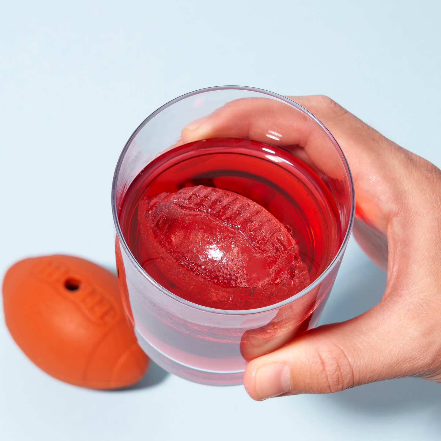 Hand holding a glass of red liquid with a football-shaped ice cube inside, next to the football shaped ice cube mold.
