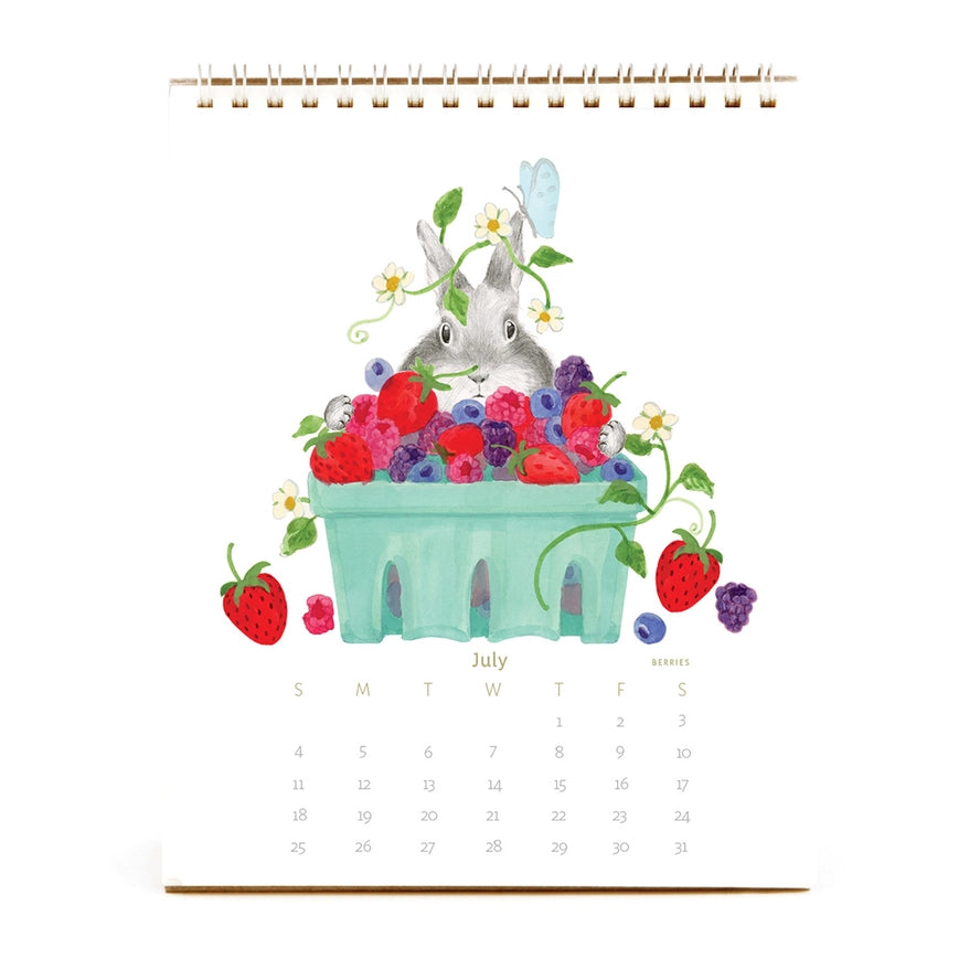 A spiral-bound desk calendar page featuring a watercolor illustration of a gray rabbit sitting in a mint green basket filled with red strawberries, blueberries, and raspberries, with the word July and a small monthly calendar grid below.