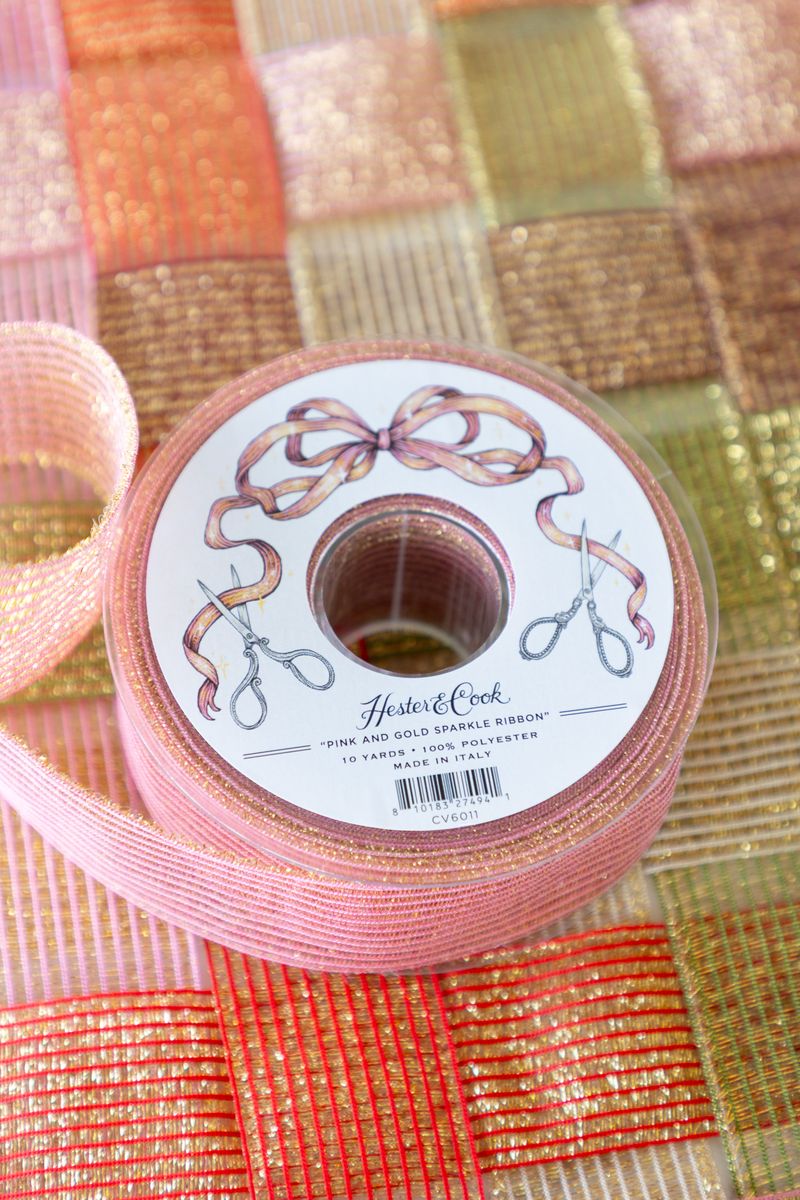 A spool of sparkly pink and gold ribbon on a surface of woven sparkle ribbon in five different colors. 
