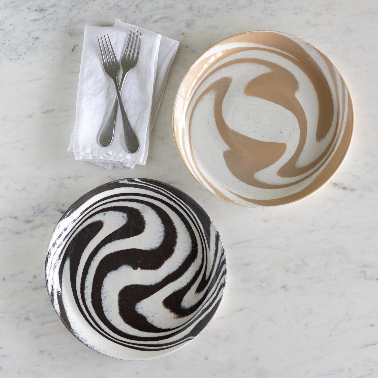 Two ceramic plates with abstract designs on a marble surface with a napkin and utensils.