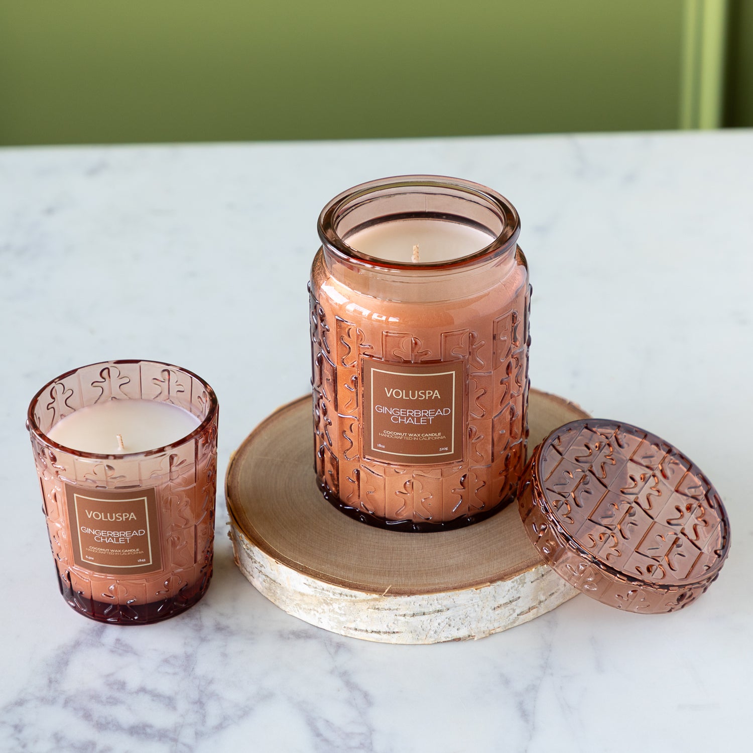 Two Voluspa candles with textured glass and wooden coaster on a marble surface.