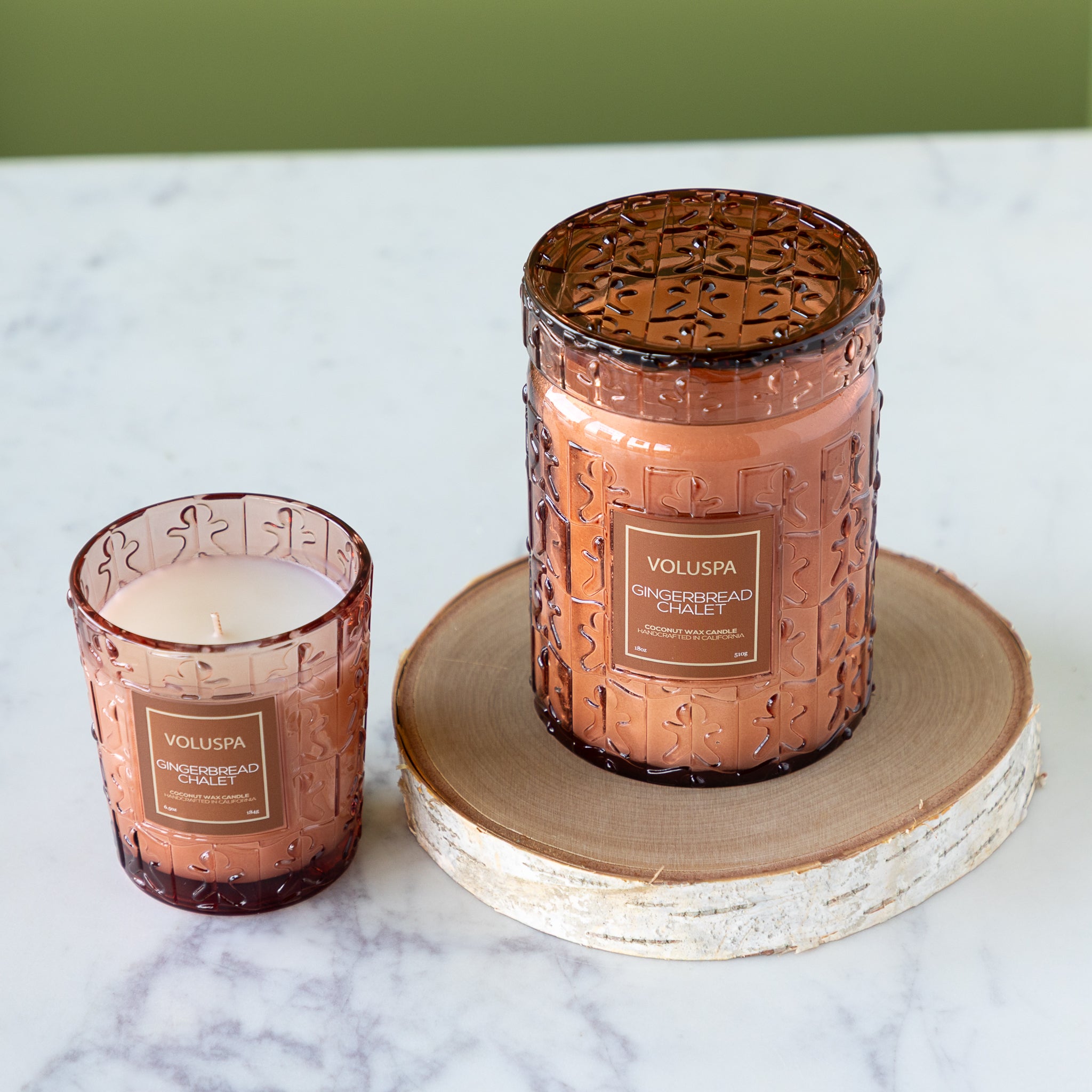 Two Voluspa candles in textured glass jars on a marble surface with a green background.