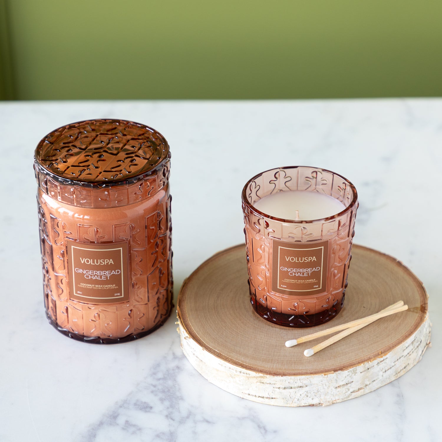 Two embossed glass candles on a wooden coaster with a green background.