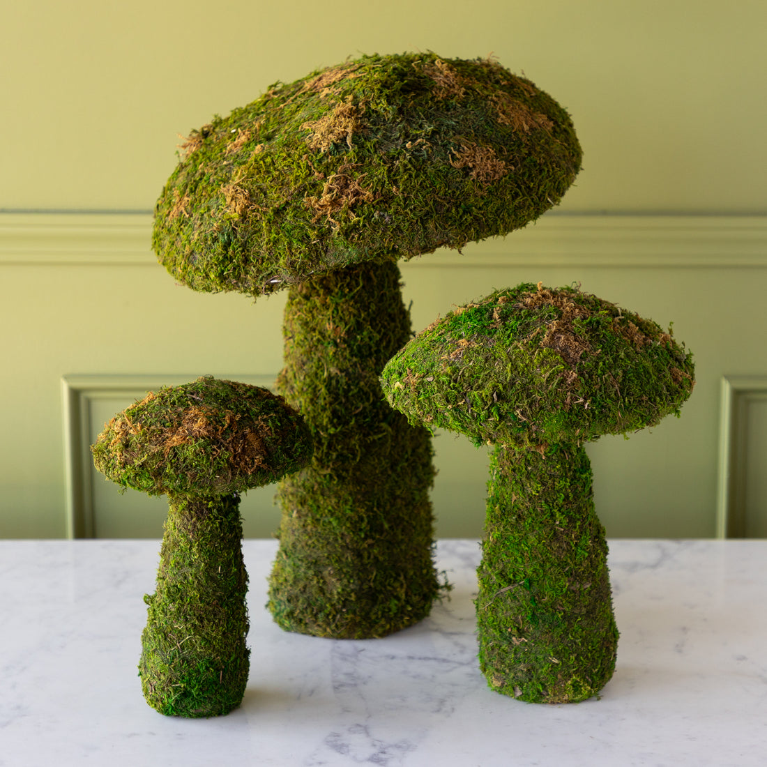 Three moss-covered mushroom planters on a marble surface with a green wall background.