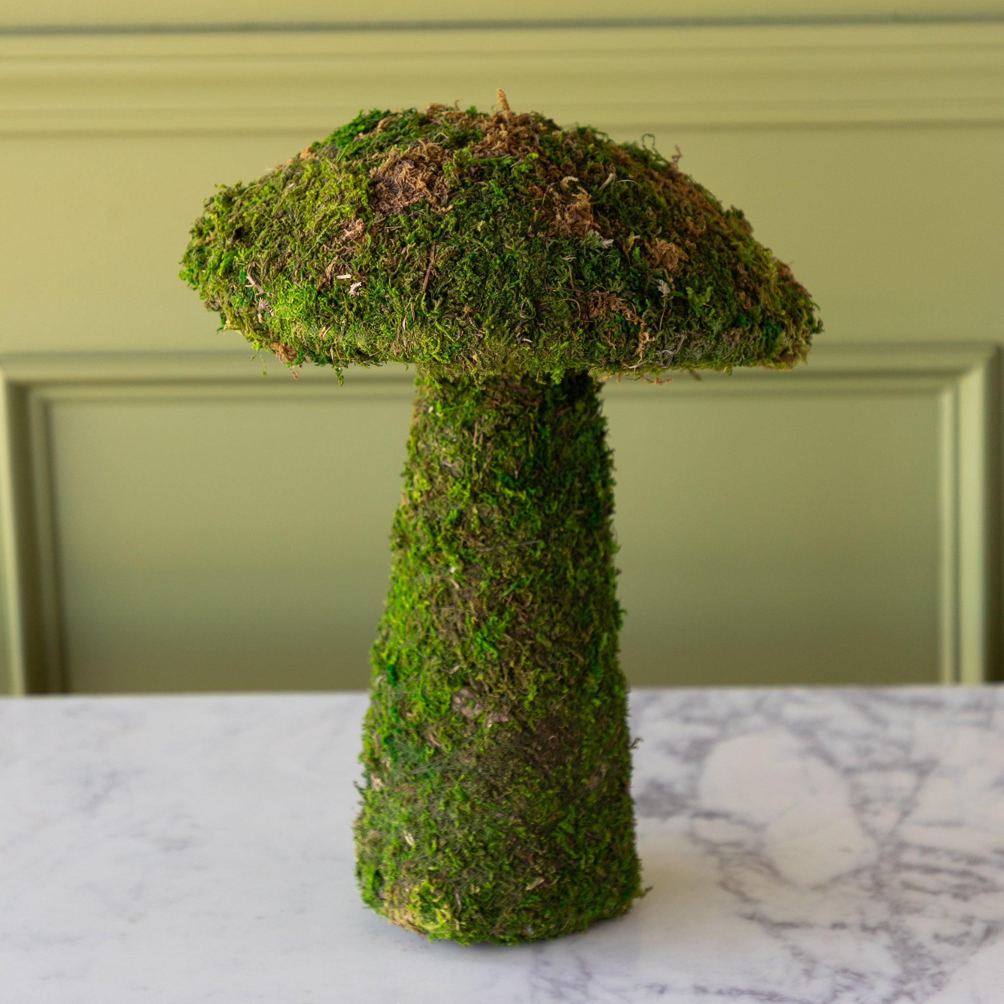 Moss-covered mushroom planter on a marble surface with a green wall background.