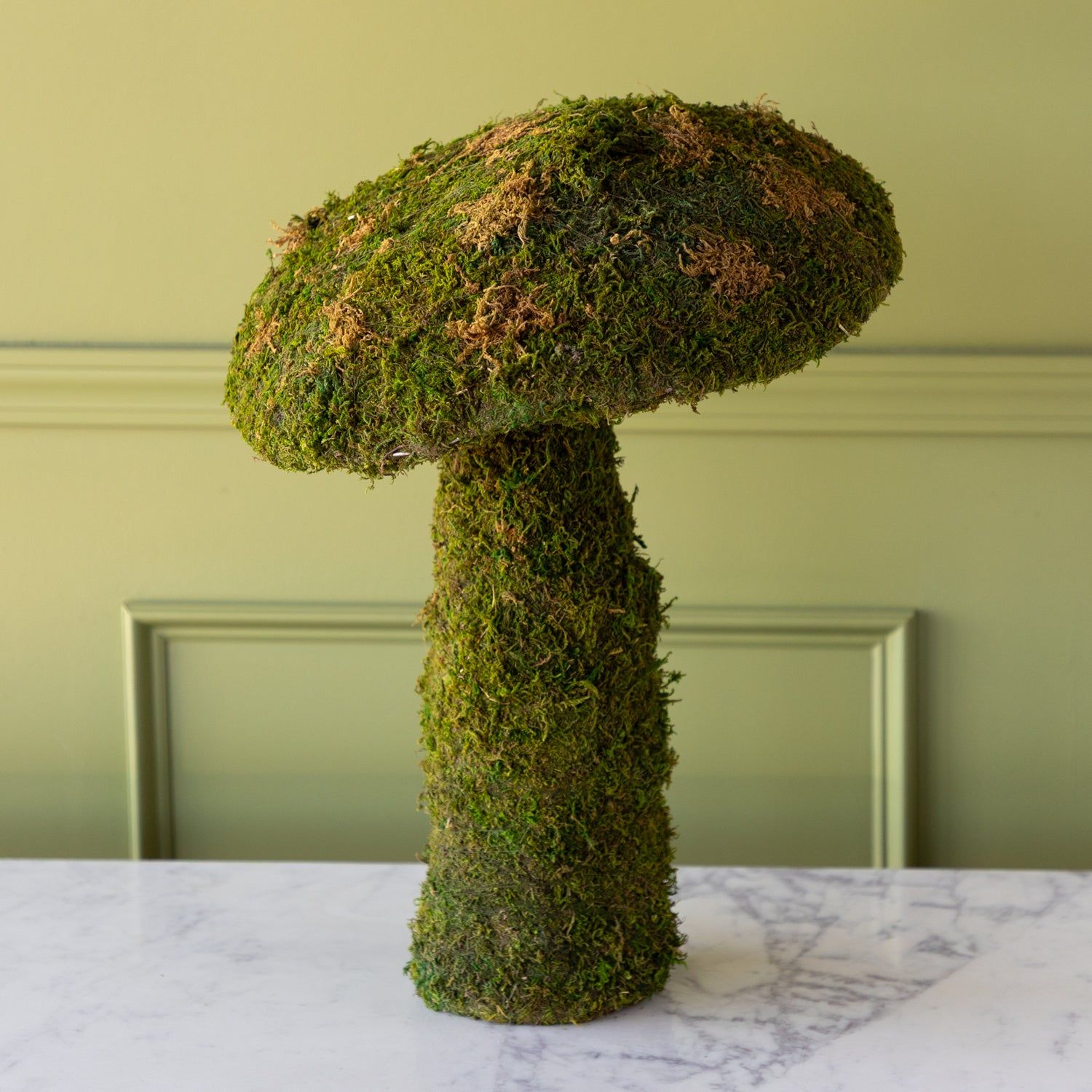 Moss-covered mushroom planter on a marble surface with a green wall background.