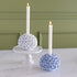 Two candle holders with white candles on a marble surface against a green wall.
