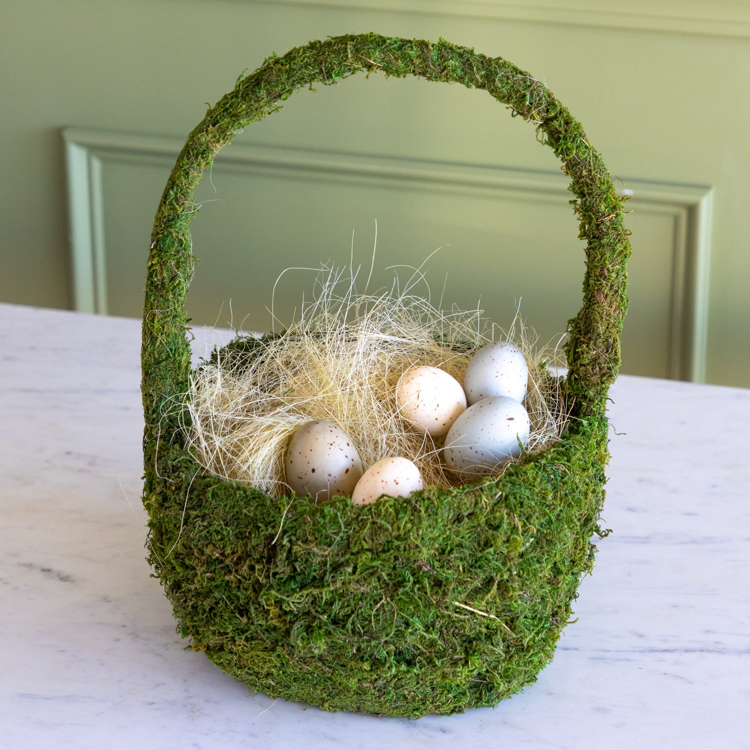 Moss-covered basket with decorative eggs on a marble surface.
