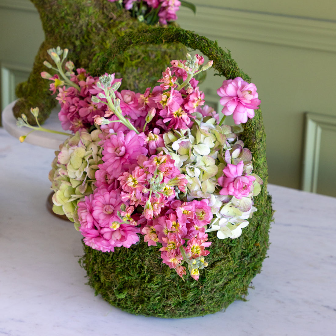 Decorative flower arrangement in a moss-wrapped basket on a white surface.