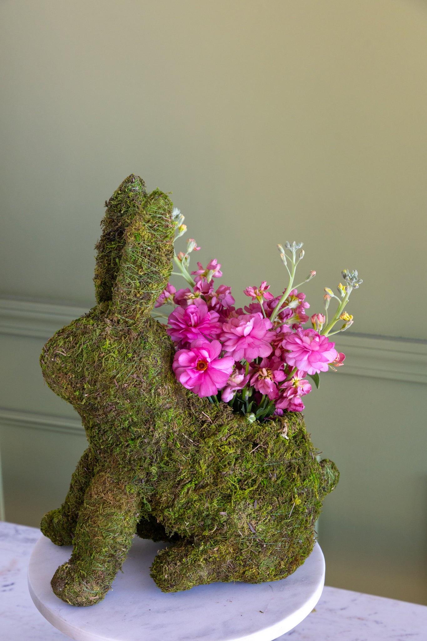 Moss-covered rabbit-shaped planter with pink flowers on a white surface against a beige wall.