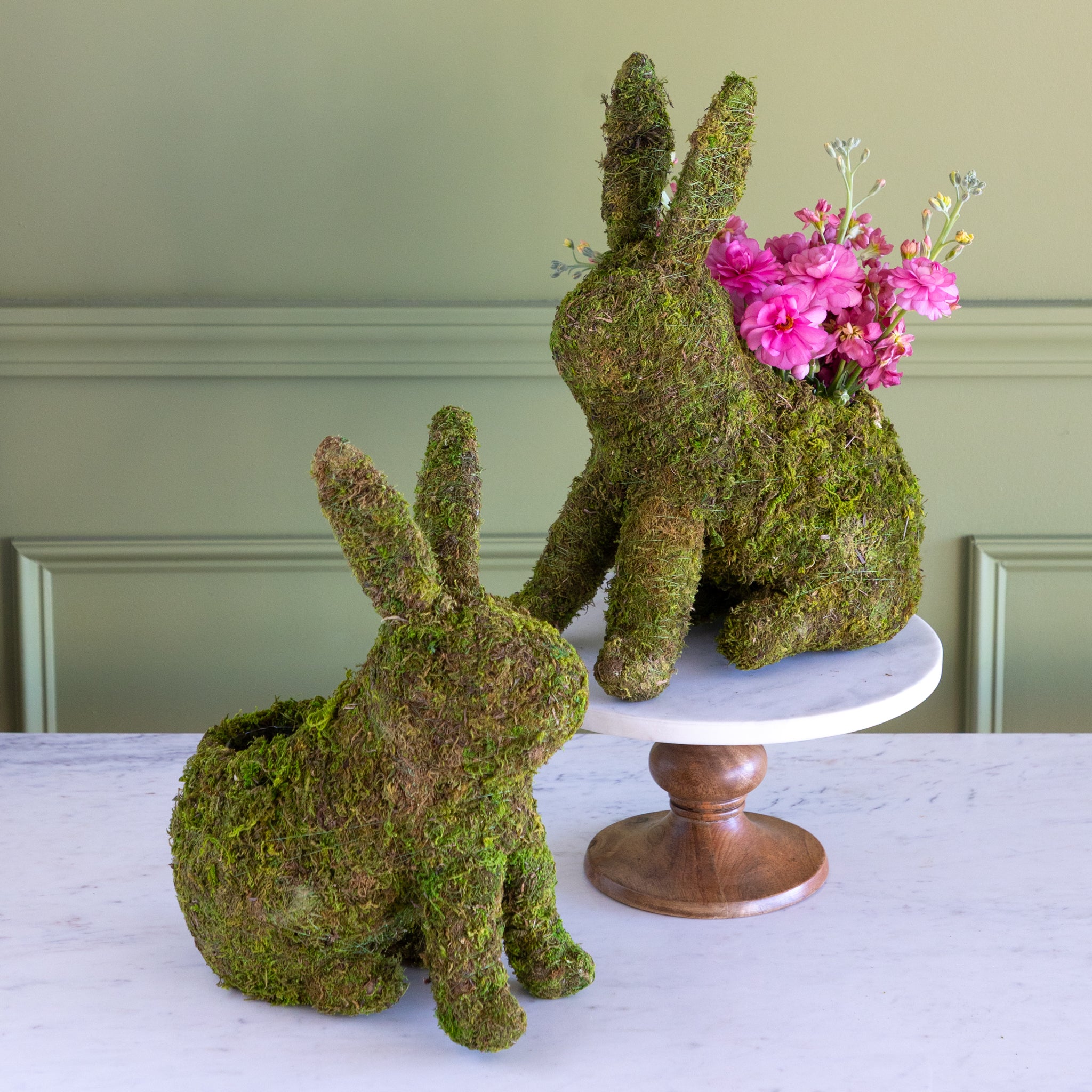 Two moss-covered bunny-shaped planters with one holding pink flowers on a white surface.