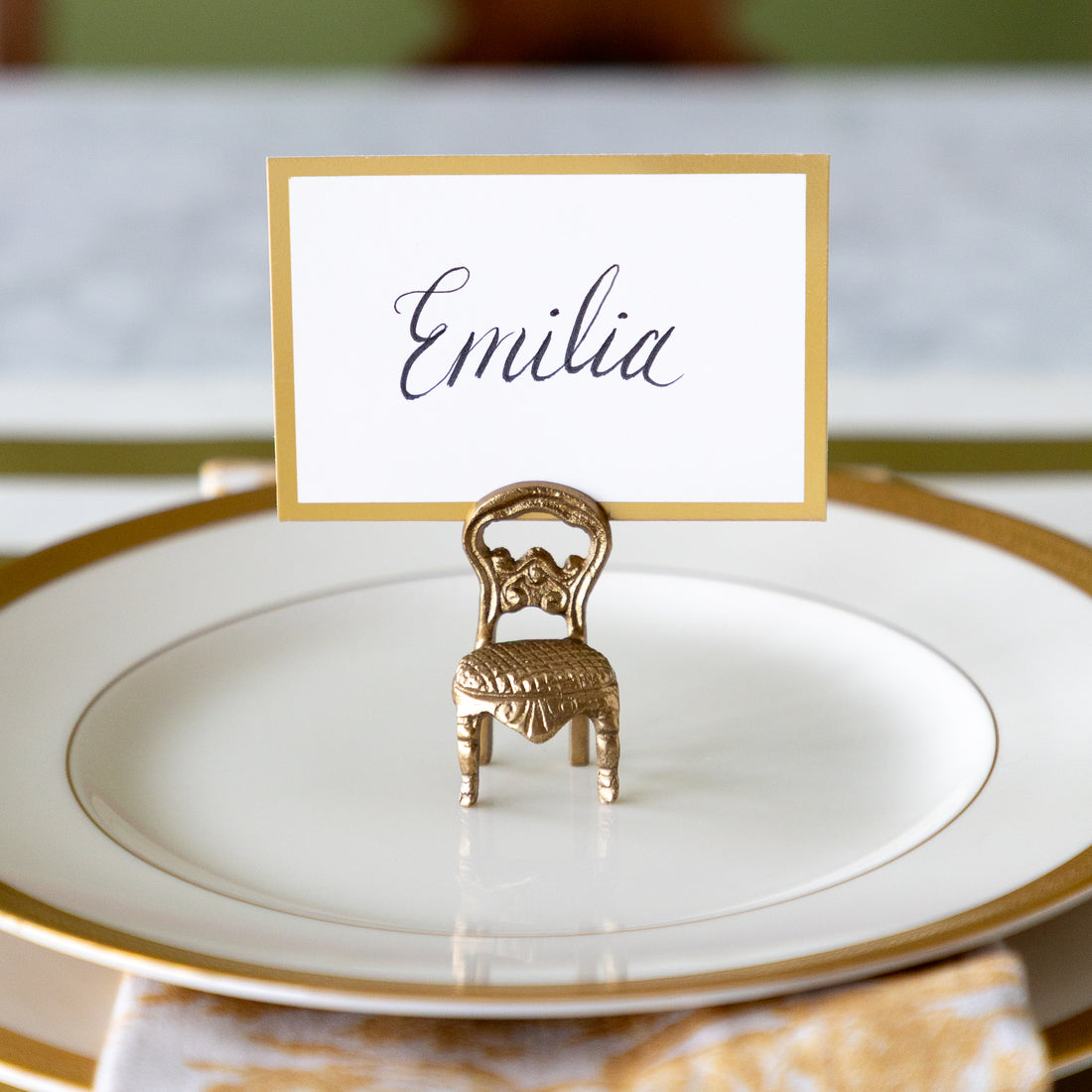 A gold miniature chair place card holder holding a white card with the name &