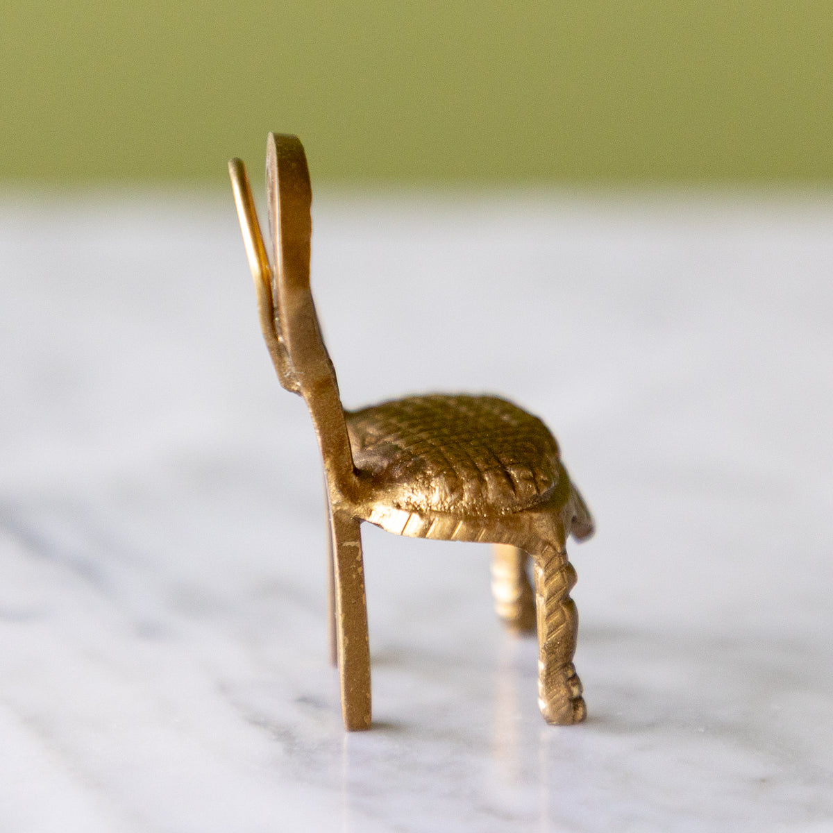 A miniature gold-colored metal chair figurine with a textured woven seat and twisted legs, standing on a white marble surface with a soft-focus olive green background.