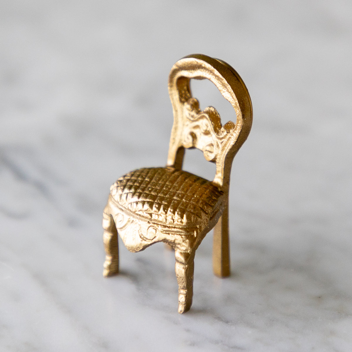 A miniature gold-colored chair figurine with a woven-texture seat and ornate backrest, displayed on a light gray marble surface.