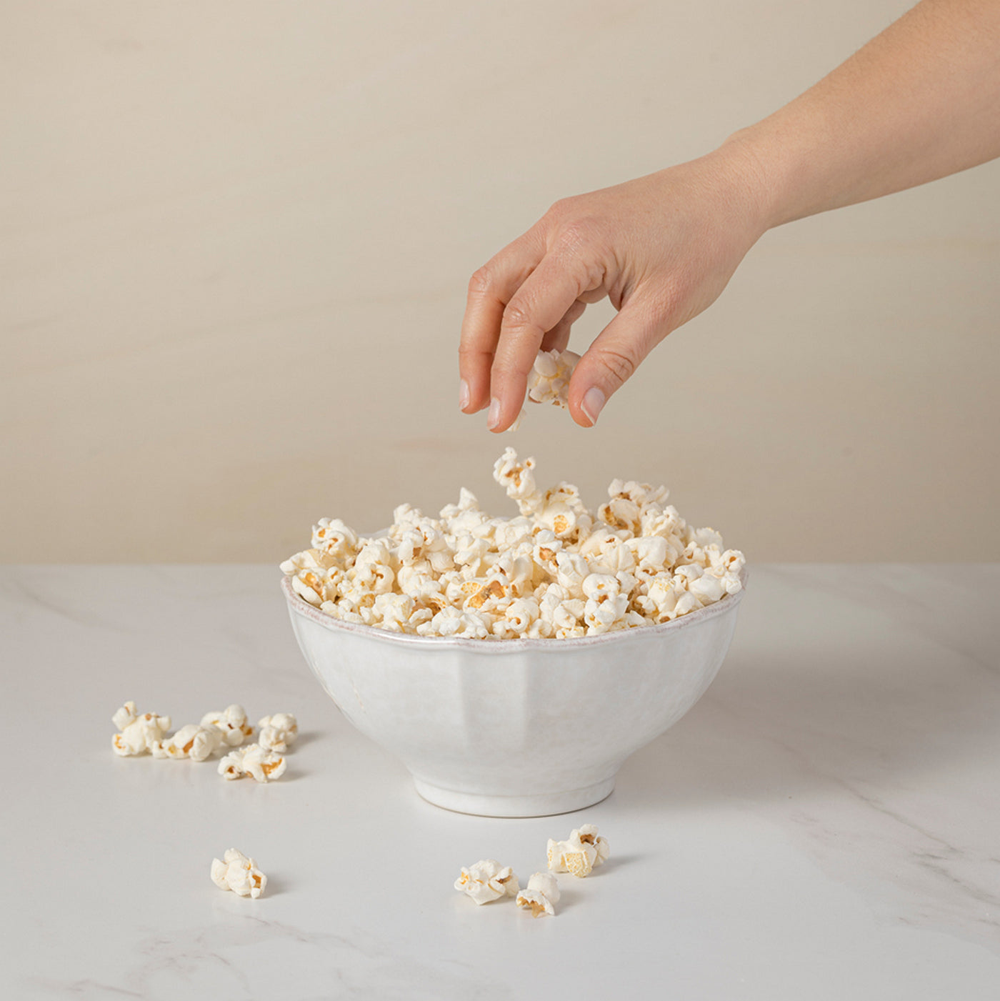Hand adding popcorn to a bowl on a light surface with a neutral background
