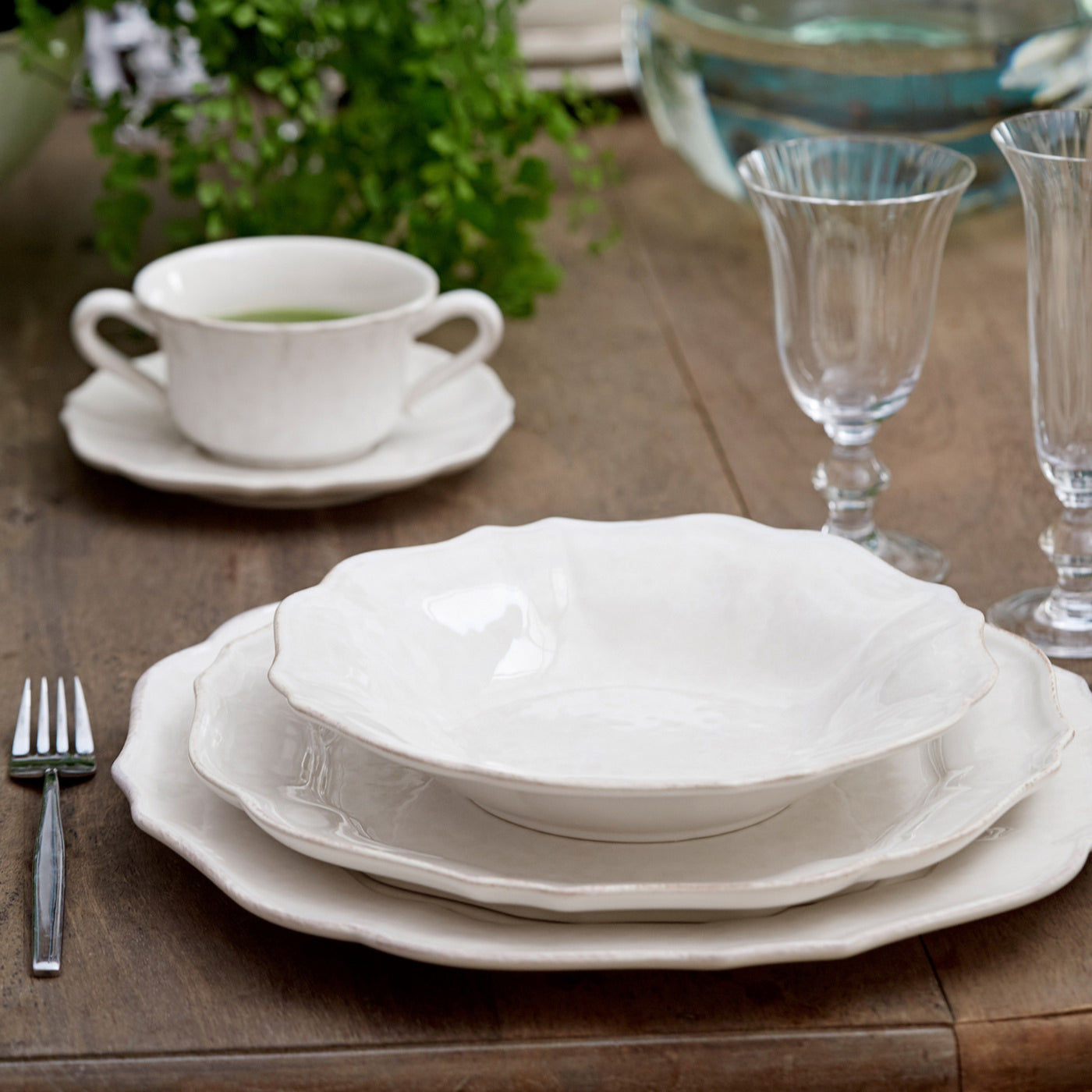 Set of white ceramic plates on a wooden table with a cup, saucer, and glasses.