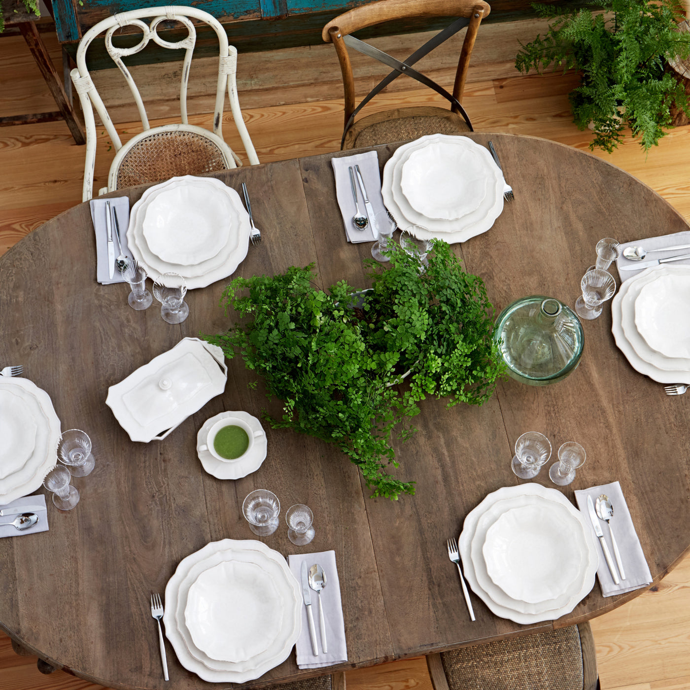 Dining table set with white plates, silverware, and greenery on a wooden floor.