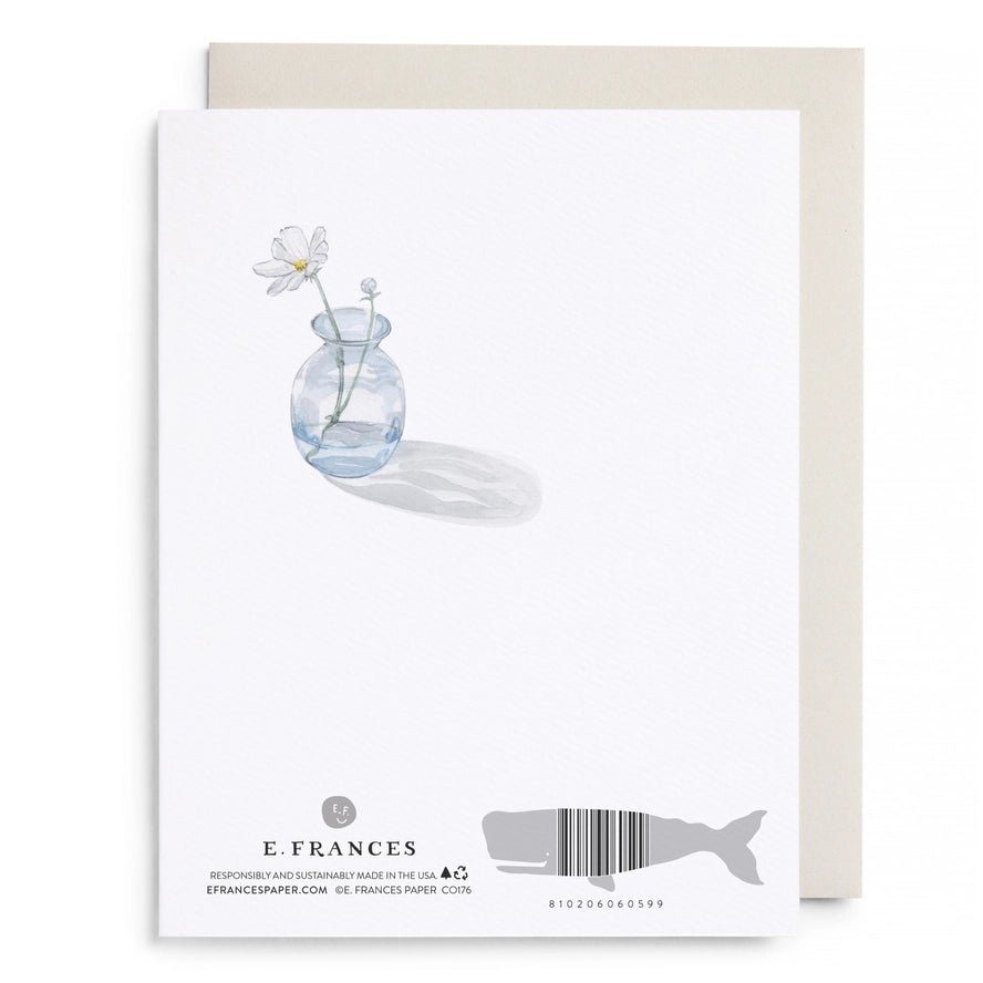 A white stationery card with a watercolor-style illustration of a small glass vase holding white flowers, shown with a beige envelope behind it on a light gray background. The card is branded E.