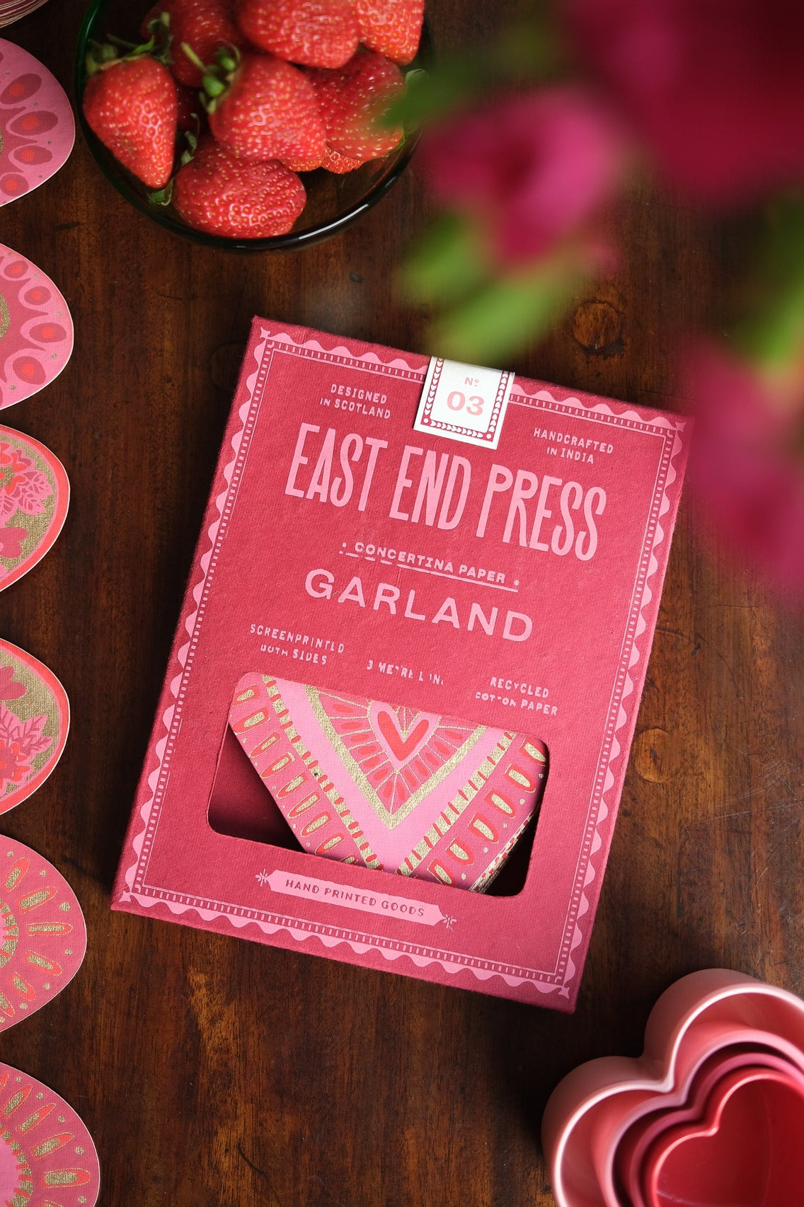 Pink packaging of East End Press Garland on a wooden surface with strawberries and flowers in the background.