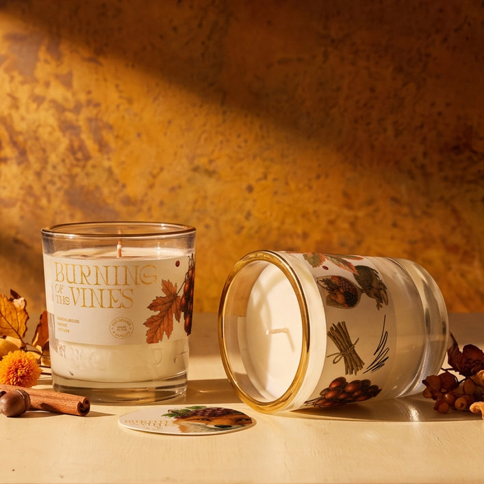 Two glass jar candles with white wax and botanical illustrations are styled on a tabletop with autumn props. One candle stands upright with its label reading &