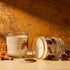 Two glass jar candles with white wax and botanical illustrations are styled on a tabletop with autumn props. One candle stands upright with its label reading &