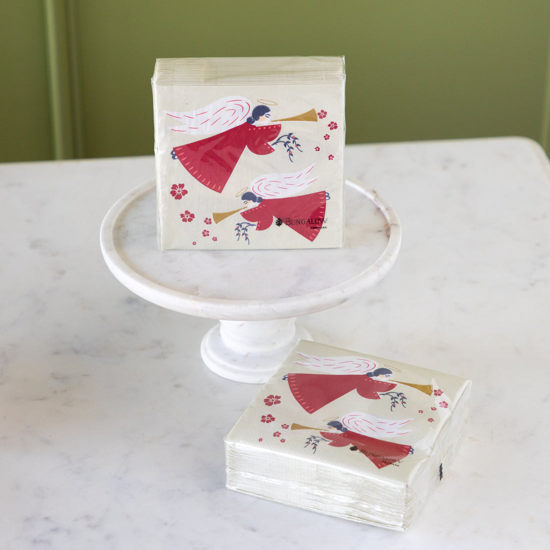 A stack of decorative paper napkins with a red angel illustration sits on a white pedestal and on a marble table. The napkin design features a red-winged angel with a gold trumpet and small red flowers on a cream background.