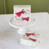 A stack of decorative paper napkins with a red angel illustration sits on a white pedestal and on a marble table. The napkin design features a red-winged angel with a gold trumpet and small red flowers on a cream background.