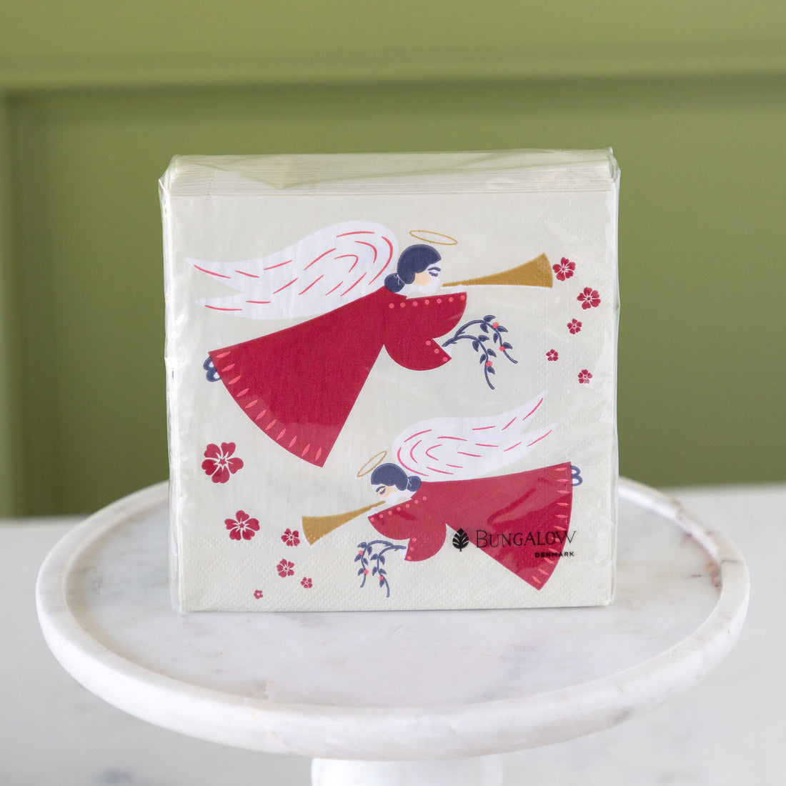 A packaged set of napkins featuring a festive illustration of two angels in red robes playing brass instruments, with small pink flowers, displayed on a white marble pedestal against a muted green wall.