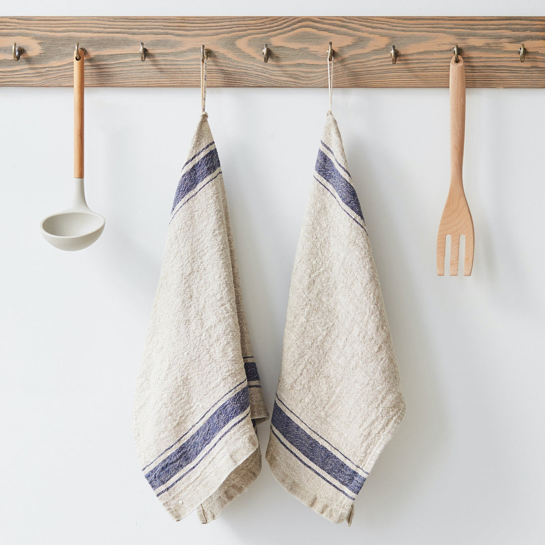 Two beige towels with blue stripes hanging on a wooden rack with kitchen utensils.