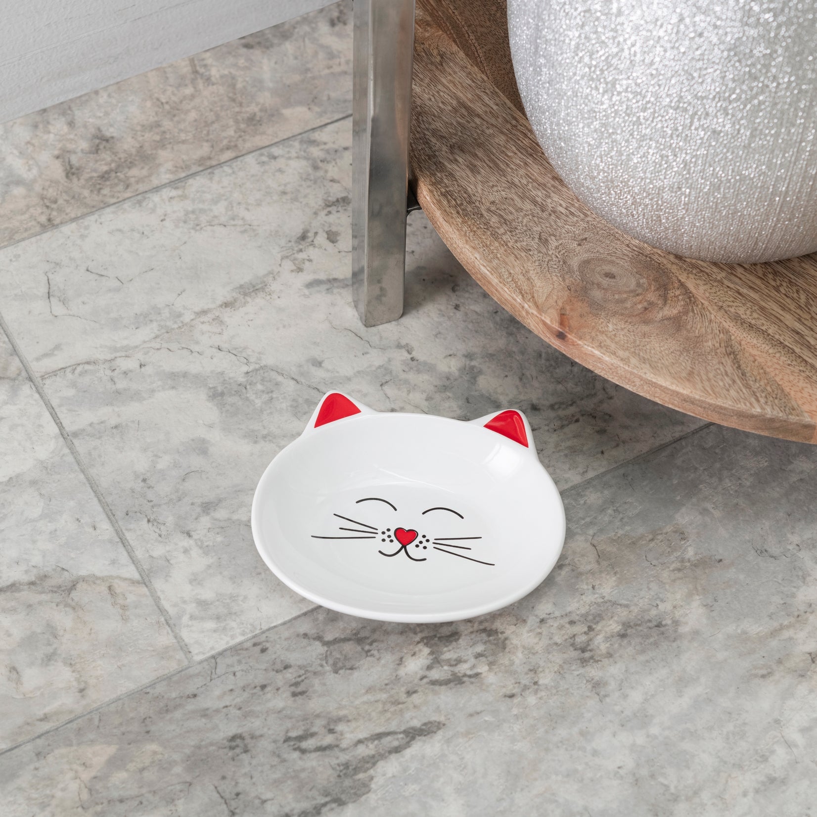 A small white cat-shaped dish with red ears and a black-and-red face design, placed on a gray marble floor near a wooden stool and a white textured vase.