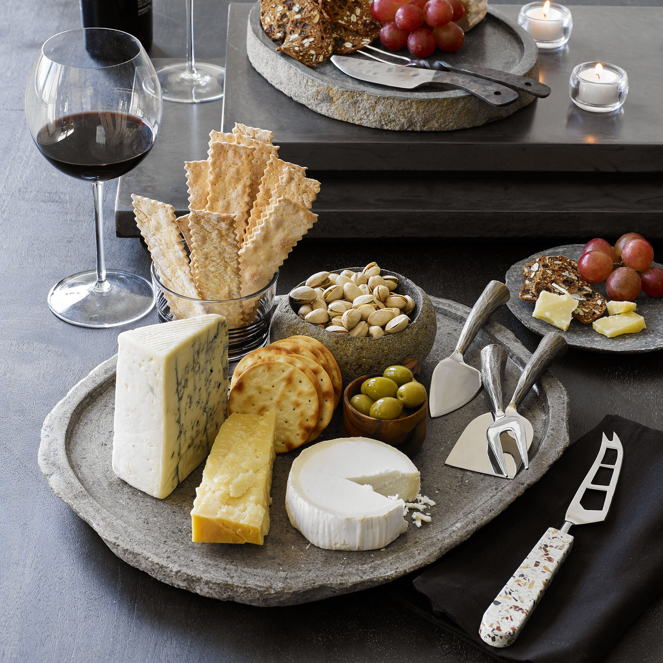 A styled charcuterie and cheese serving setup on a dark tabletop. A round stone platter holds assorted cheeses, crackers, nuts, olives, grapes, and cheese knives.