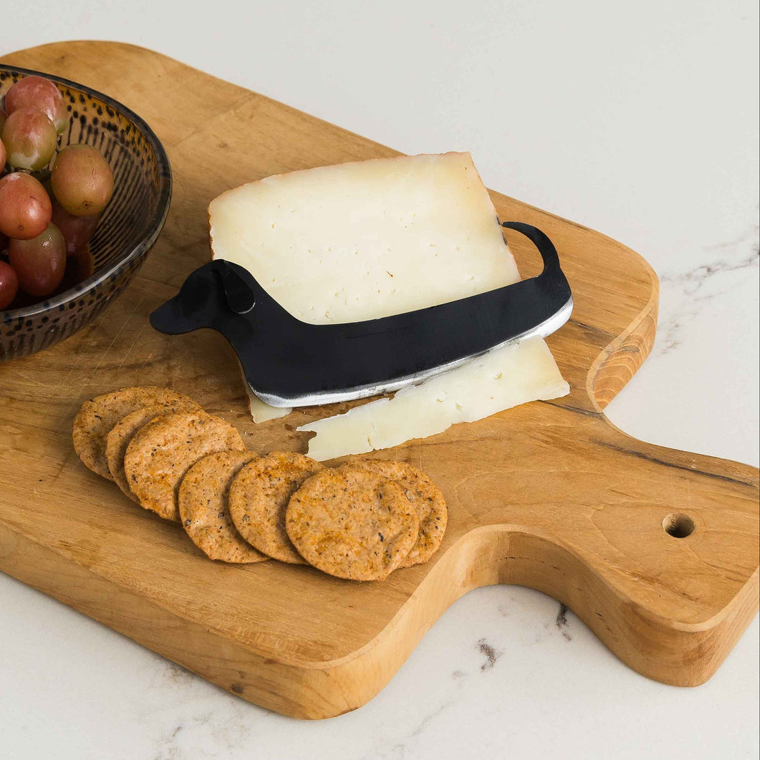 A cheese knife in the shape of a dachshund cutting a piece of cheese on a cutting board with crackers. 