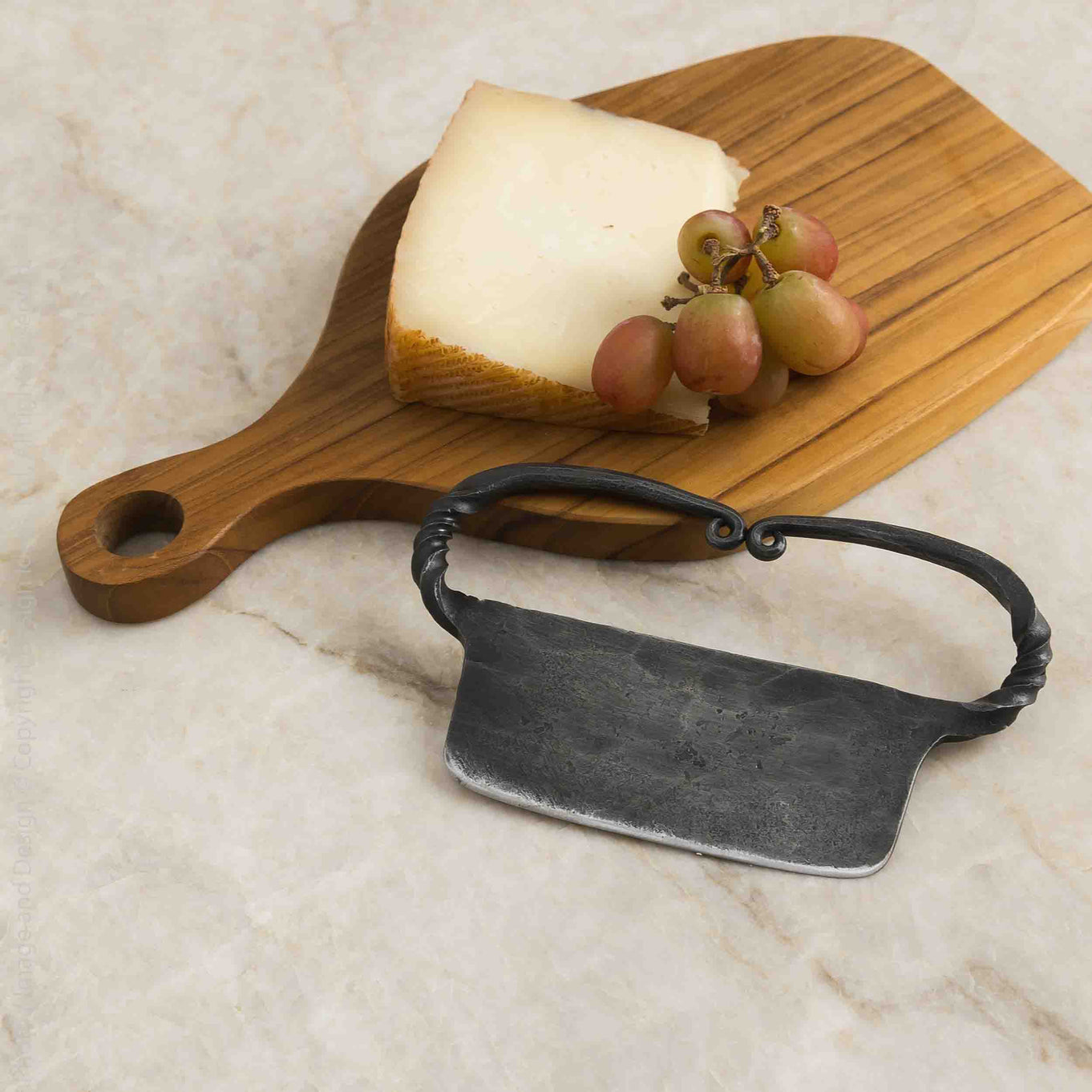 Wooden cutting board with cheese and grapes, accompanied by a black metal cheese knife on a marble surface.