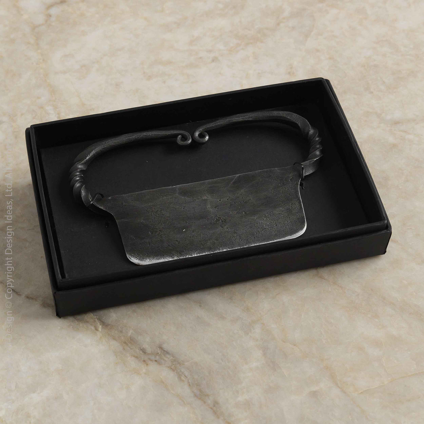 A dark metal cheese knife with a textured, hammered finish and a decorative handle, presented inside a black gift box on a beige marble surface.