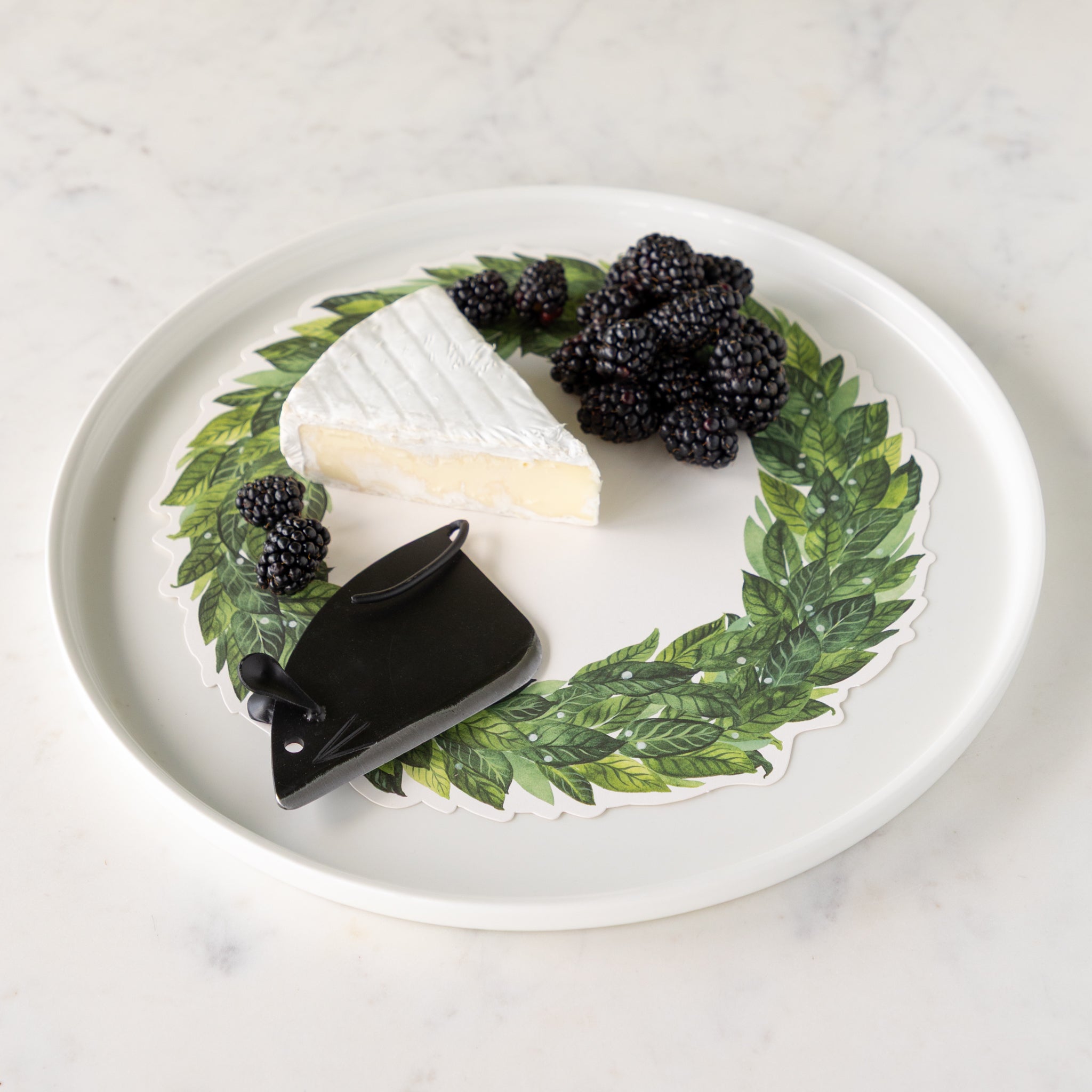 White plate with a wedge of cheese, blackberries, and a black cheese knife shaped like a mouse on a marble surface.
