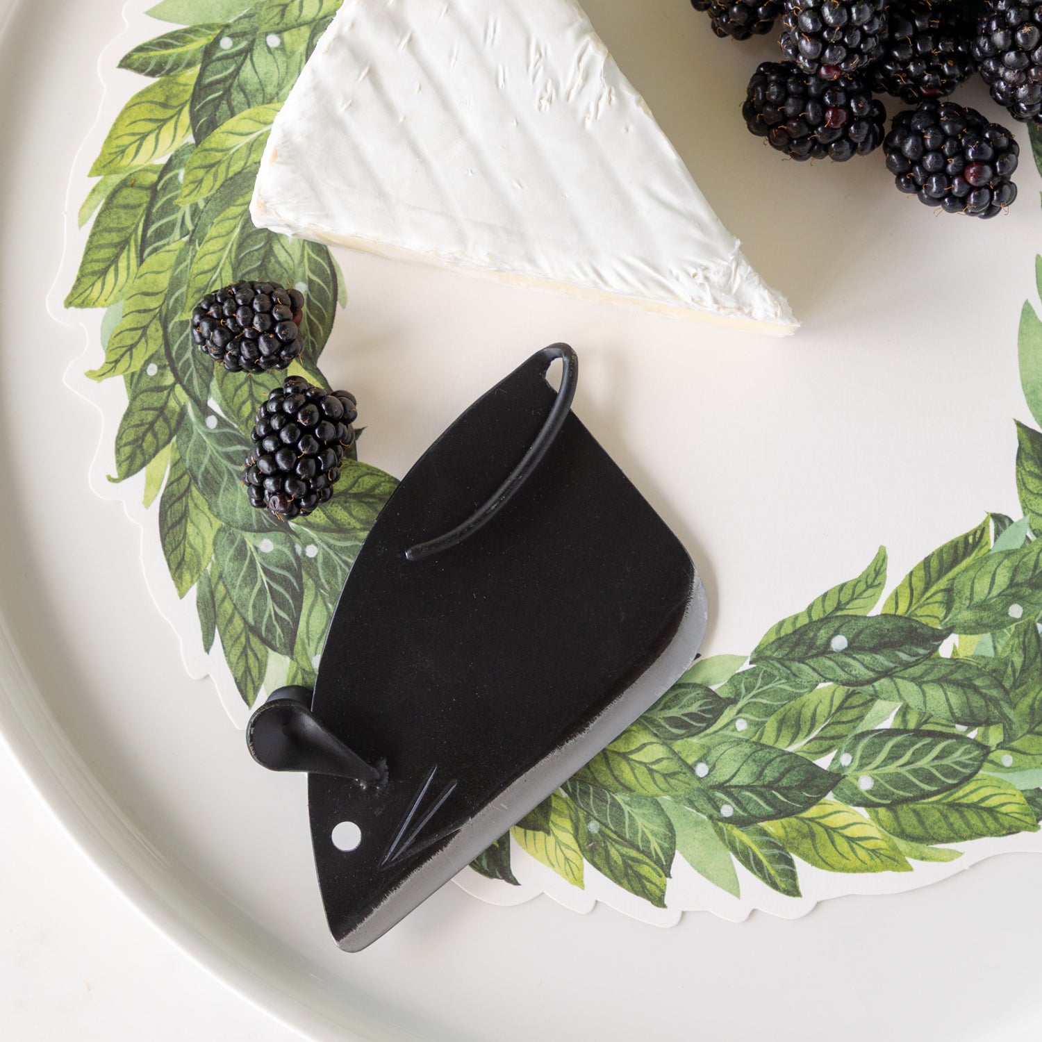 Black mouse-shaped cheese cutter on a decorative plate with the Laurel Serving Paper, blackberries and a wedge of cheese.