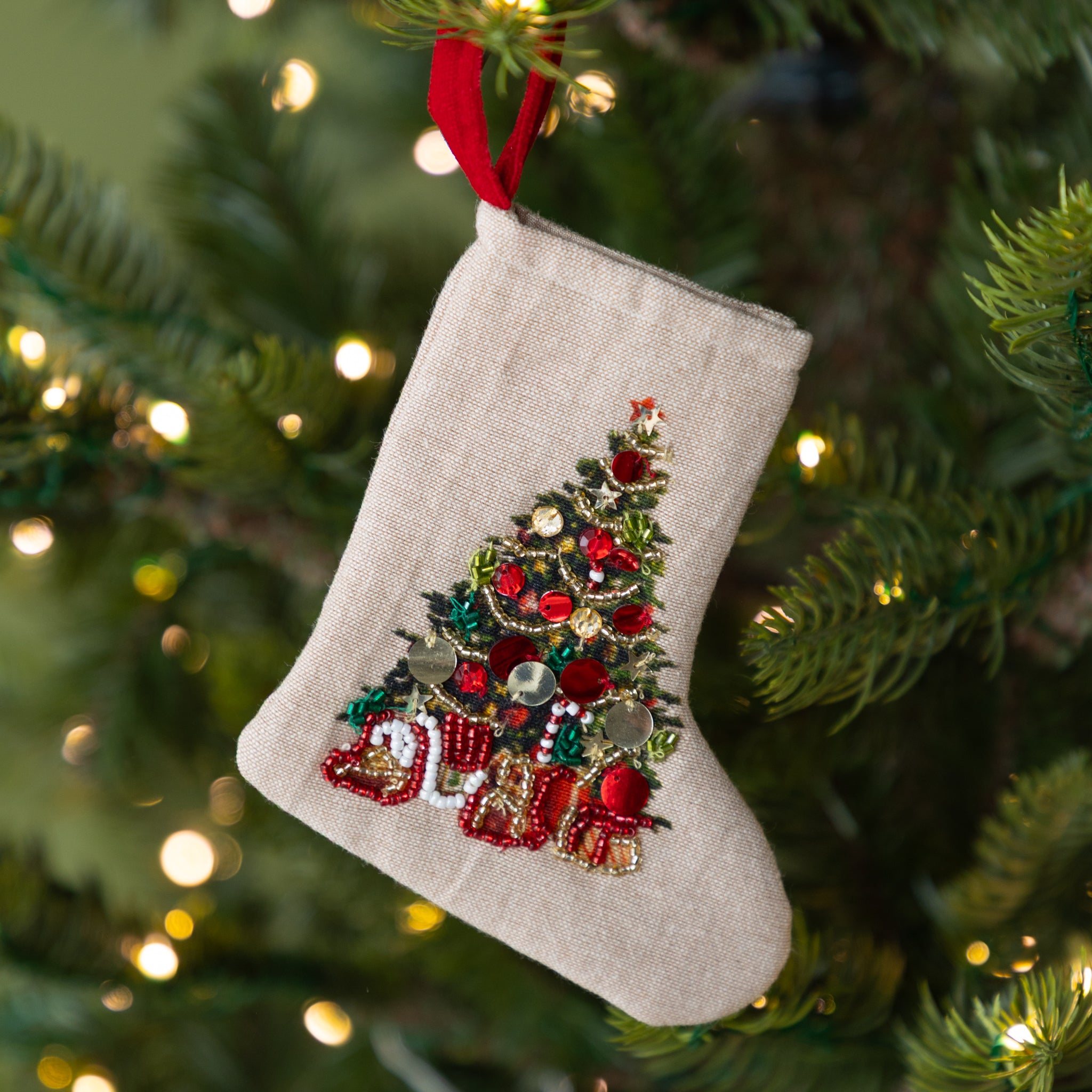 Decorative Christmas stocking with a tree and presents design, hanging on a Christmas tree by a red loop.