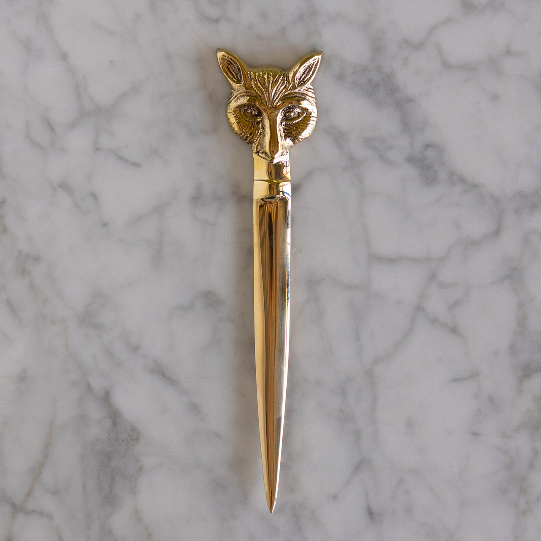 A polished gold-toned metal letter opener with an intricately detailed fox head handle, resting vertically on a white and gray veined marble surface.