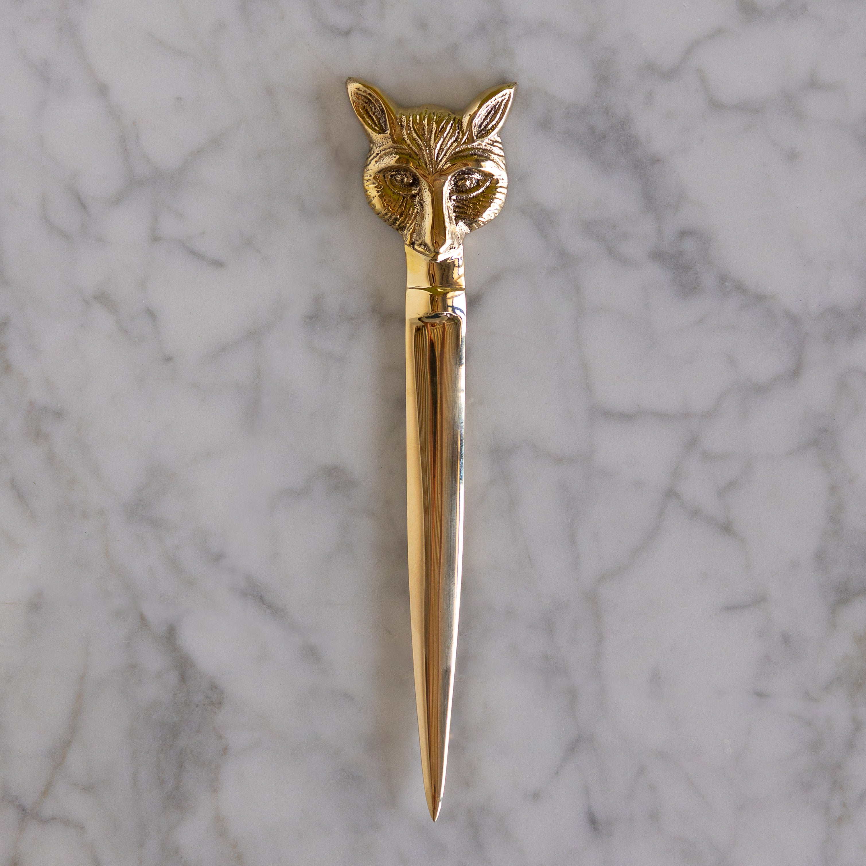 A polished gold-toned metal letter opener with an intricately detailed fox head handle, resting vertically on a white and gray veined marble surface.