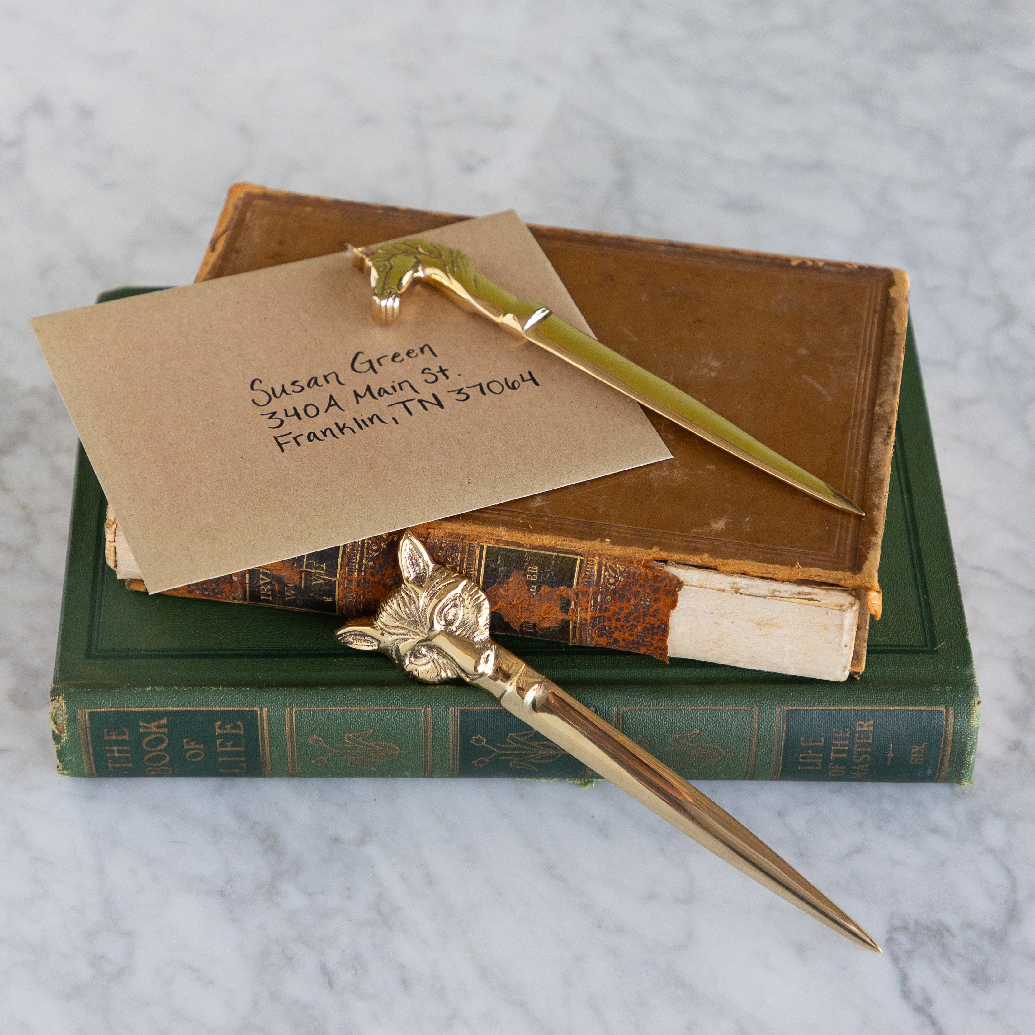 A gold-toned metal letter opener with a fox head handle rests on top of a brown vintage book, with a brown kraft paper business card placed beside it. Another green vintage book is underneath, all photographed on a light marble surface.
