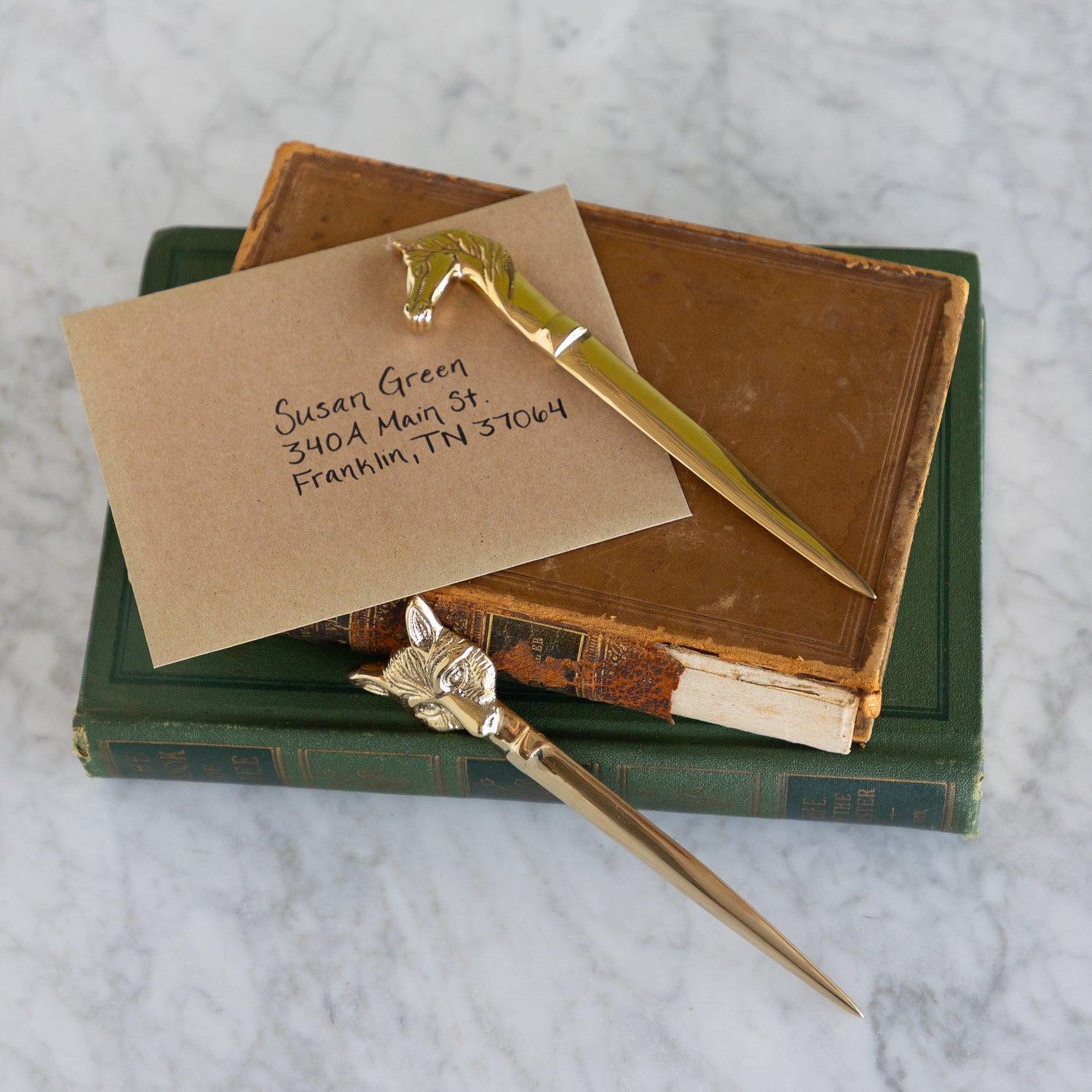 Two gold-toned metal letter opener style stationery pieces with decorative animal heads, arranged on top of an old brown book and a kraft paper card addressed by hand, on a green book and a white marble surface.