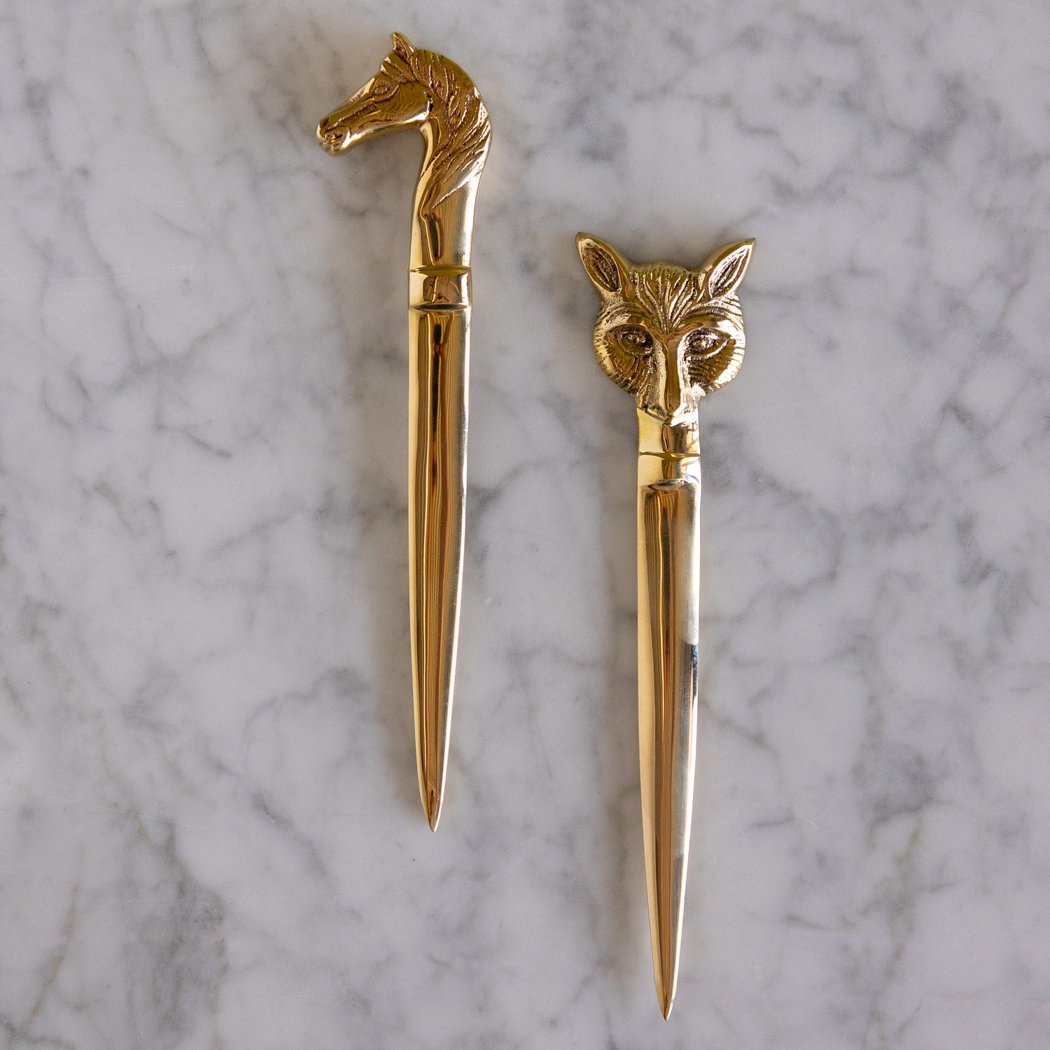 Two gold-toned letter opener style desk accessories with animal head handles, one horse-shaped and one fox-shaped, displayed on a white marble surface.