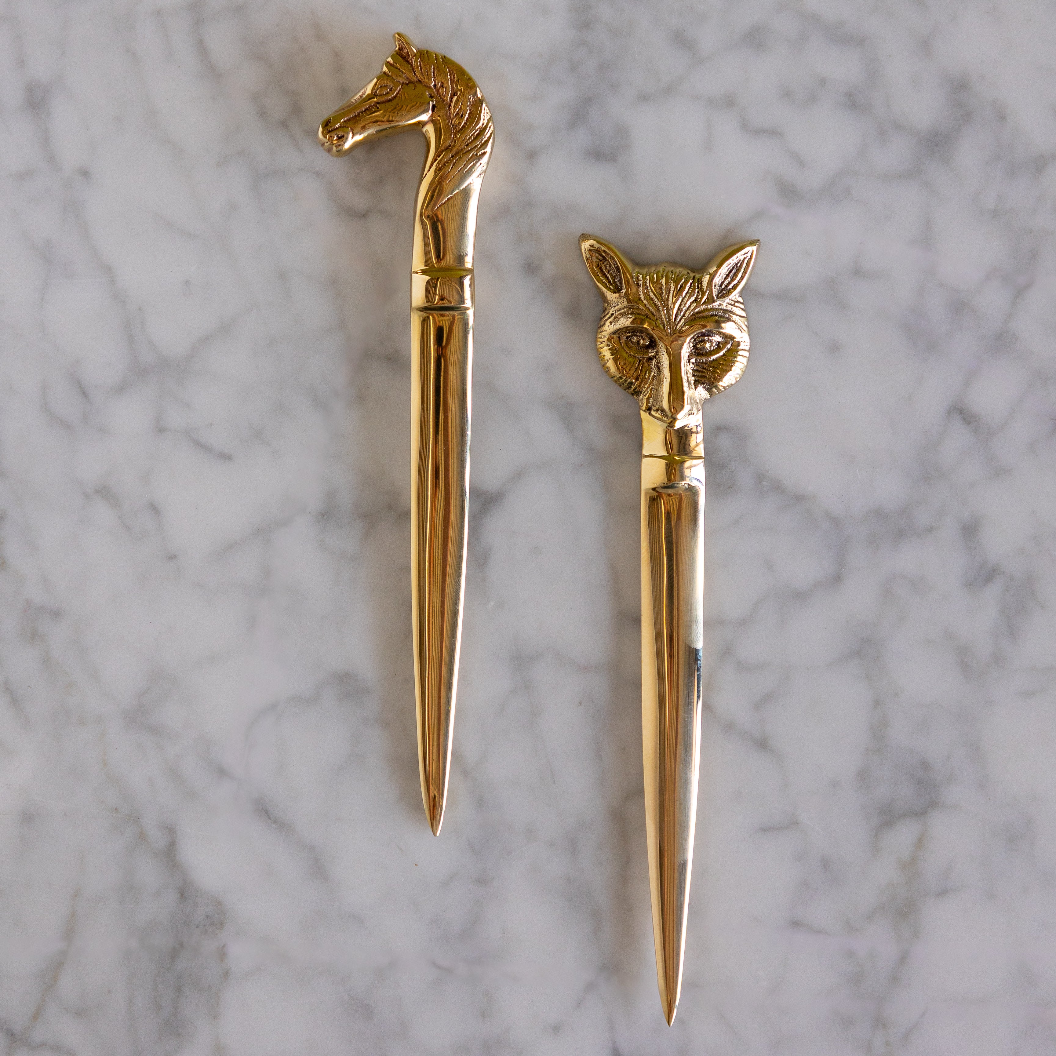 Two gold-toned letter opener style desk accessories with animal head handles, one horse-shaped and one fox-shaped, displayed on a white marble surface.